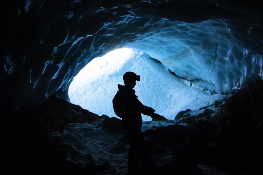 Explorador con linterna en la cabeza, de pie dentro de una cueva azulada con paredes de hielo, observando el interior del espacio subterráneo. especie