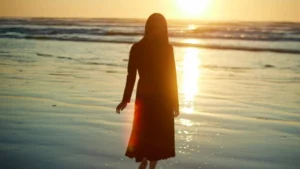 Mujer de espaldas caminando por la orilla del mar al atardecer, iluminada por la luz cálida del sol poniente.